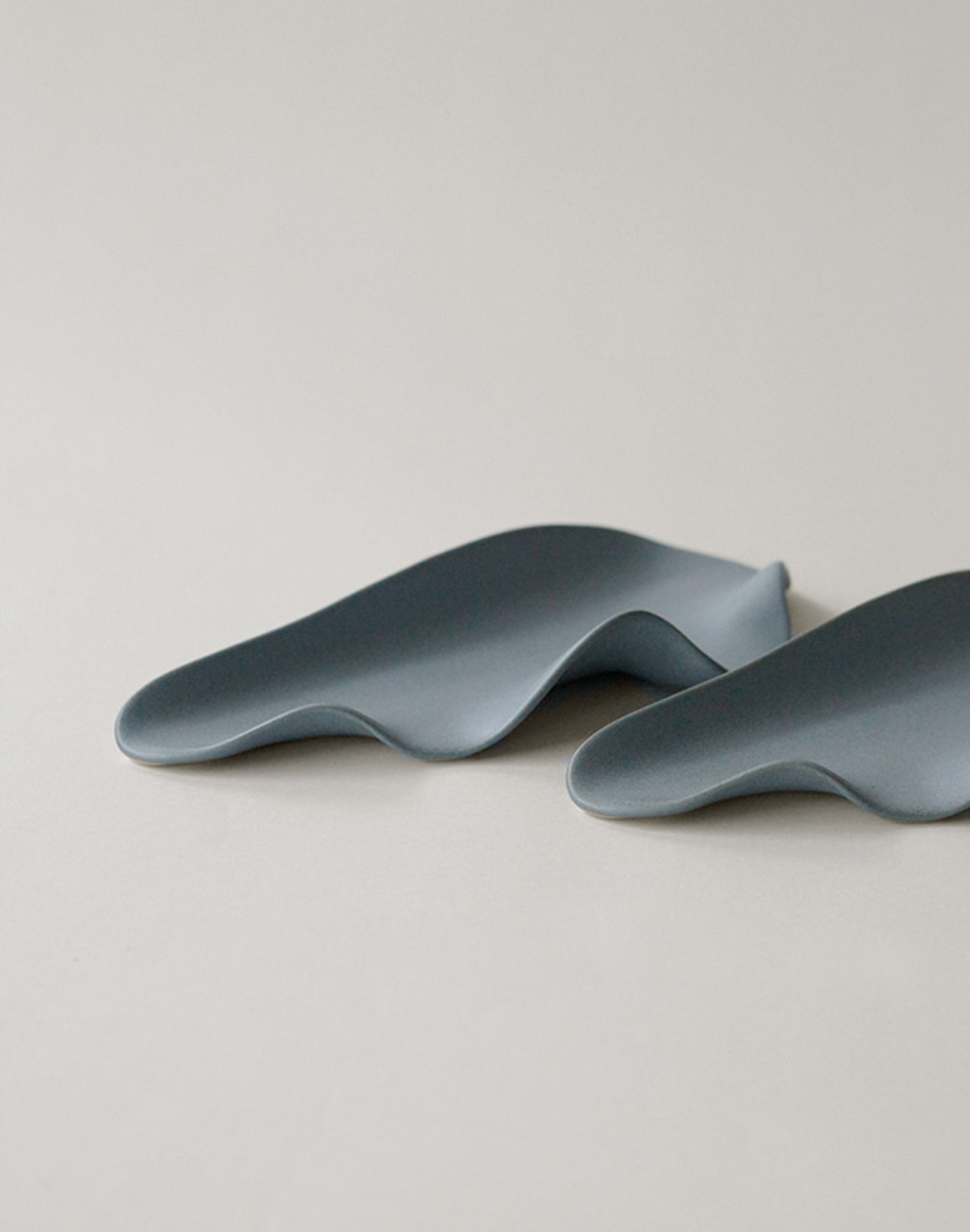 Two smooth, gray NR Ceramics HIN Leaf Plates with wavy, organic forms rest on a light gray surface—their elegant, abstract shapes evoke natural contours against a plain backdrop.