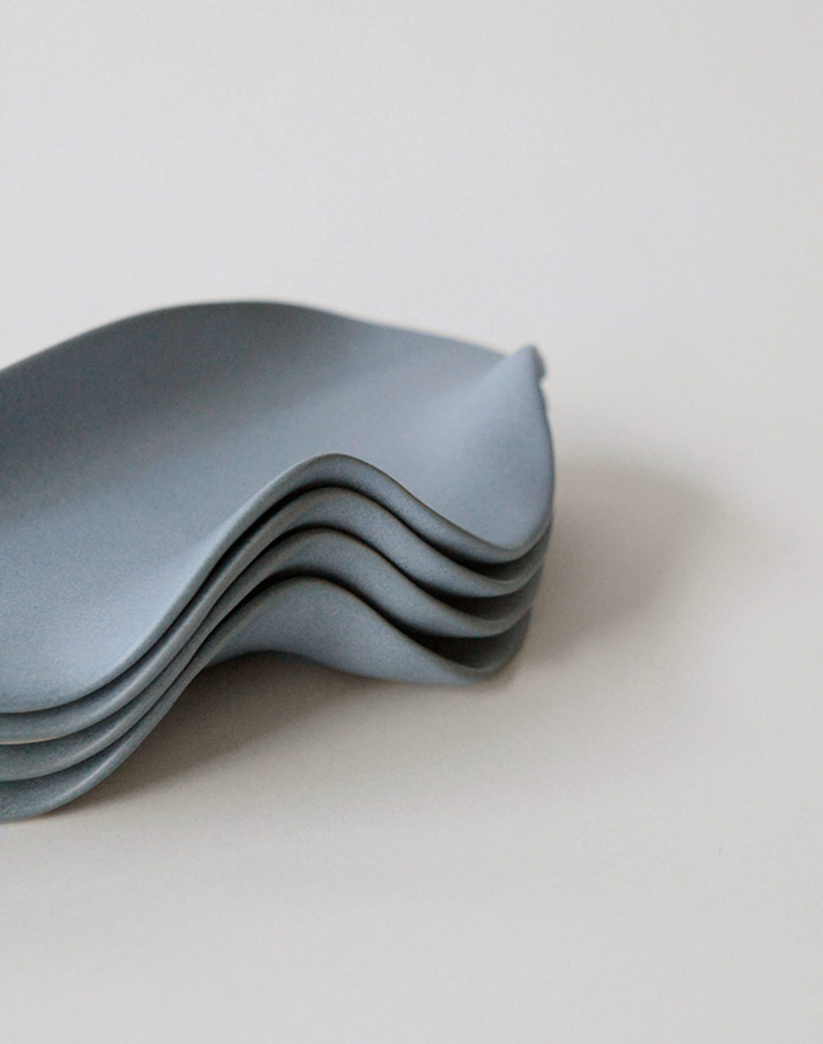 A stack of four NR Ceramics HIN Leaf Plates, featuring a minimalist wavy edge and matte grey finish, is shown on a plain background. The sculptural plates by NR Ceramics offer an organic shape for versatile use and stylish presentation.