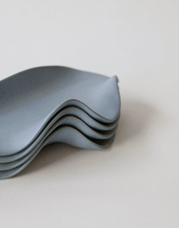 A stack of four NR Ceramics HIN Leaf Plates, featuring a minimalist wavy edge and matte grey finish, is shown on a plain background. The sculptural plates by NR Ceramics offer an organic shape for versatile use and stylish presentation.