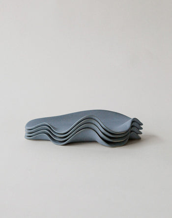 Five flat, matte blue-gray NR Ceramics HIN Leaf Plates with wavy, irregular edges are stacked on a plain background, highlighting NR Ceramics’ sculptural tableware that is both stylish and multi-functional.