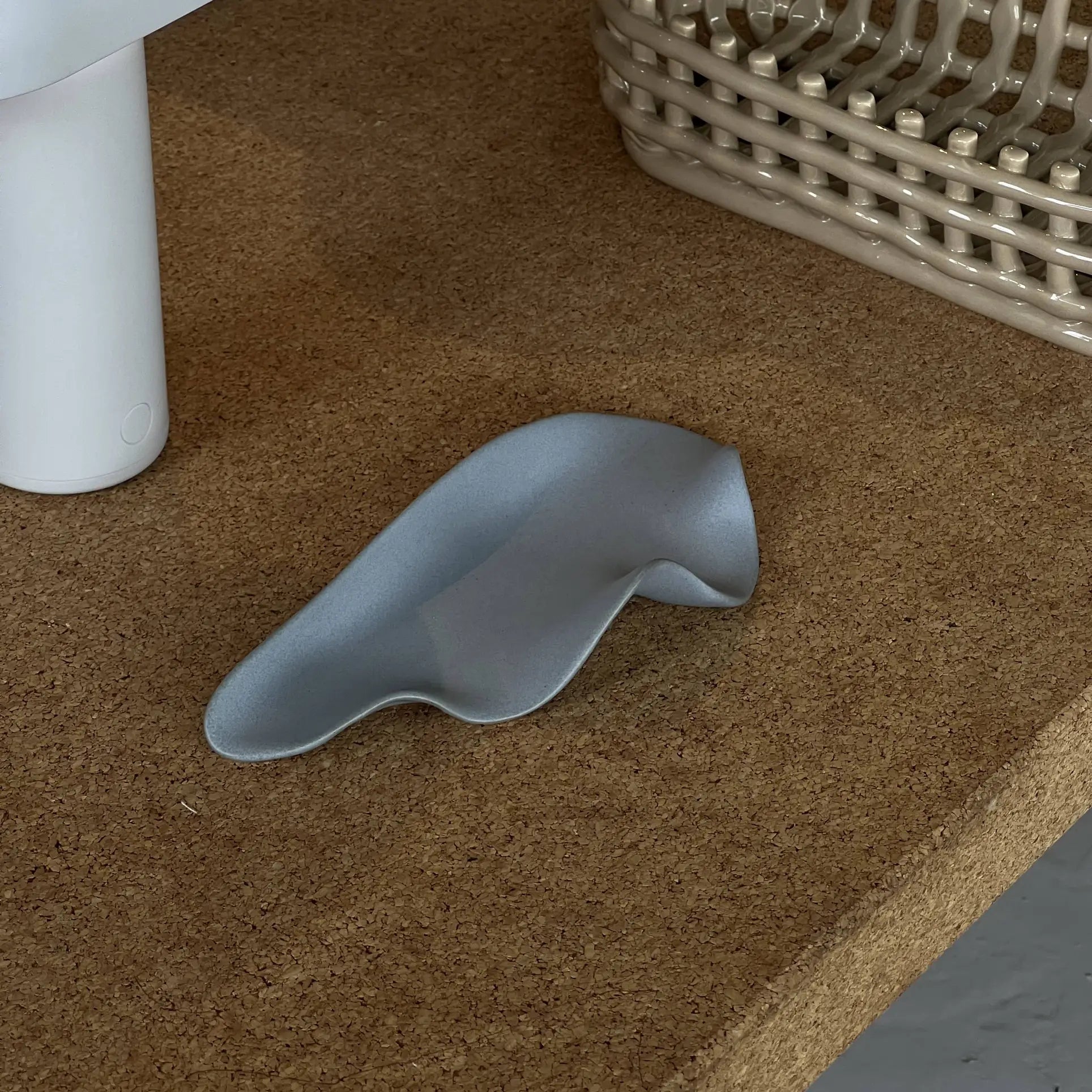 The NR Ceramics HIN Leaf Plate by NR Ceramics, in gray and abstract design, rests on a cork surface beside a white cylinder and beige basket, adding multifunctional tableware elegance to the scene.