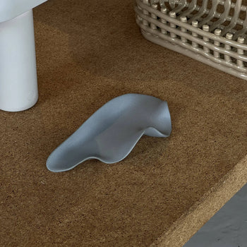 The NR Ceramics HIN Leaf Plate by NR Ceramics, in gray and abstract design, rests on a cork surface beside a white cylinder and beige basket, adding multifunctional tableware elegance to the scene.