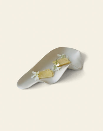 The NR Ceramics HIN Leaf Plate by NR Ceramics displays two cheeses adorned with white flowers and dill, demonstrating elegant and versatile tableware against a plain cream background.
