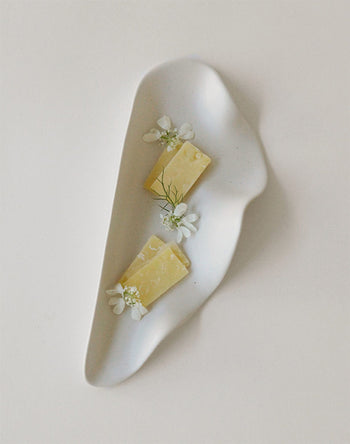 The NR Ceramics HIN Leaf Plate by NR Ceramics features two slices of yellow cheese with white flowers and green herbs, blending modern multi-functional elegance with a minimalist white background.
