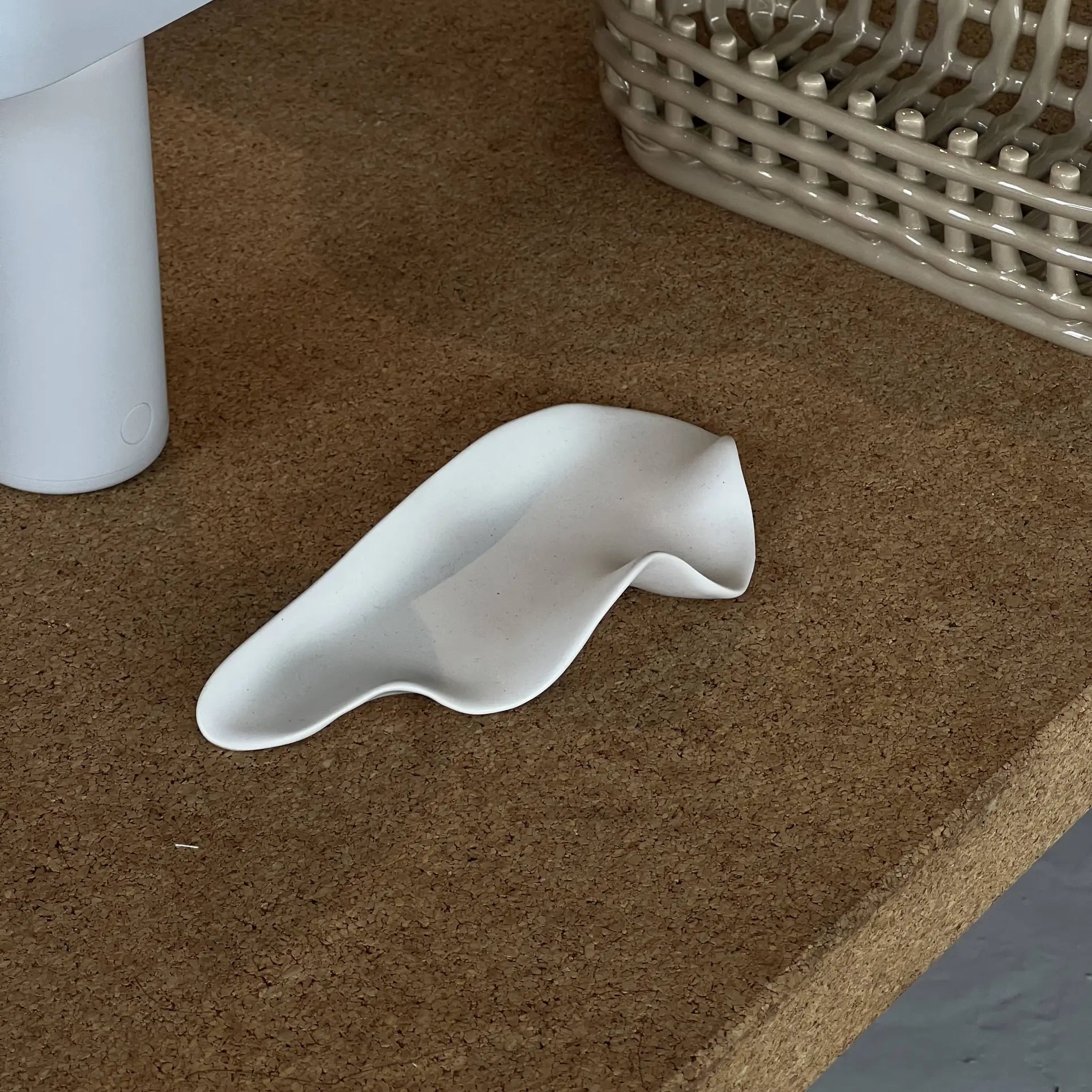 The NR Ceramics HIN Leaf Plate, a white abstract wavy ceramic dish, sits on a cork surface near a woven basket and a cylindrical white object.