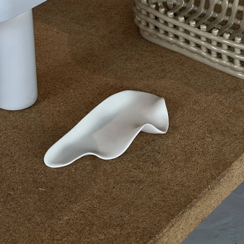 The NR Ceramics HIN Leaf Plate, a white abstract wavy ceramic dish, sits on a cork surface near a woven basket and a cylindrical white object.