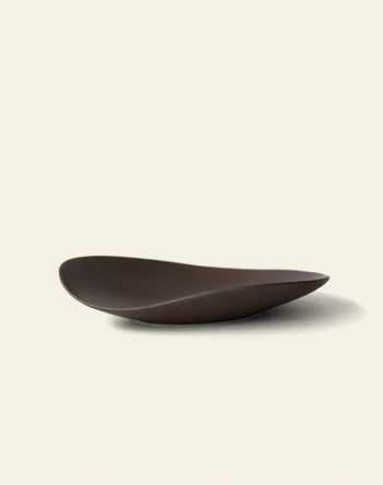 The NR Ceramics HIN Pebble Plate S is a dark brown, modern ceramic plate with an asymmetrical, smooth wavy design, photographed on a plain off-white background.