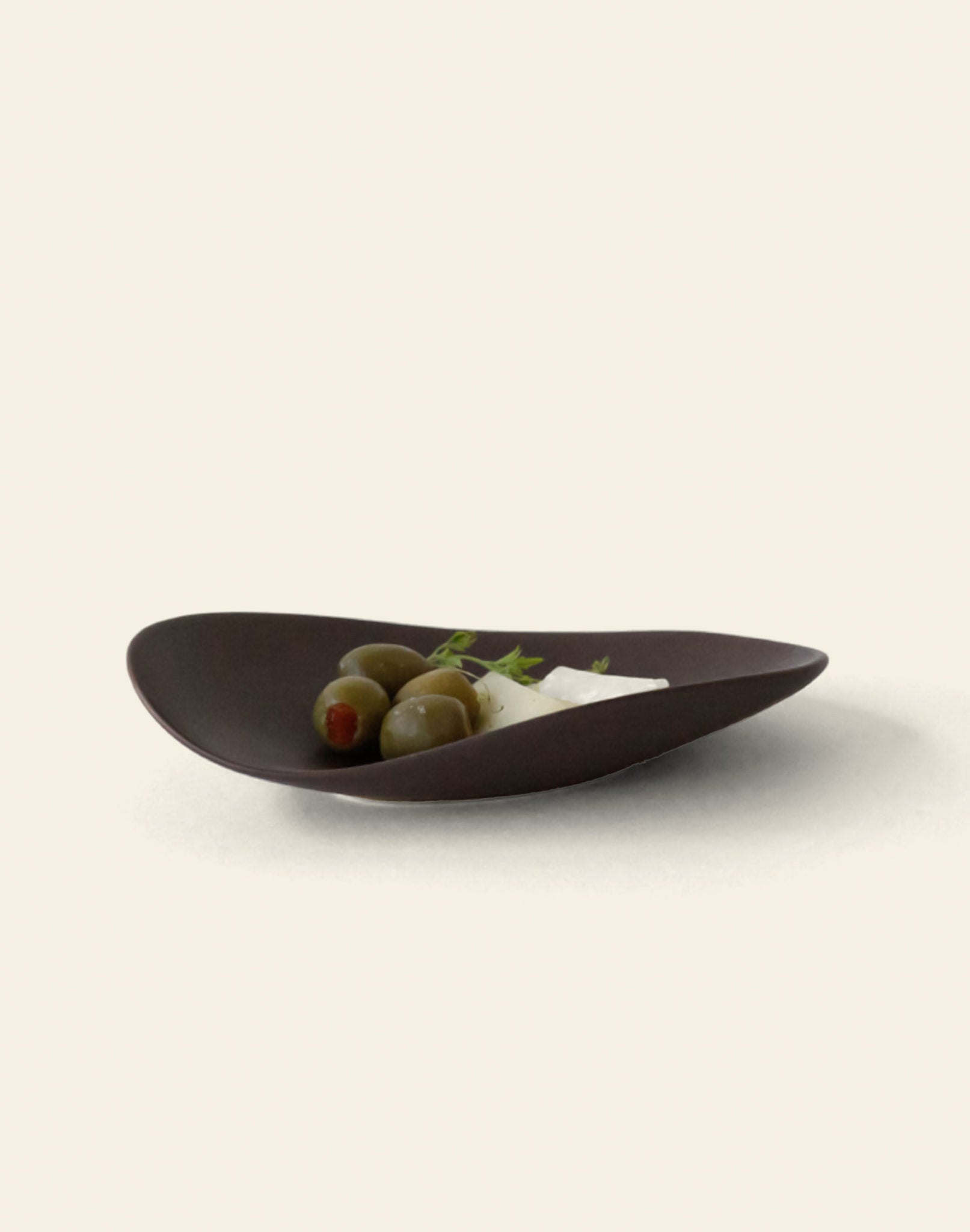 The NR Ceramics HIN Pebble Plate S, a dark, modern piece from NR Ceramics, elegantly holds three green olives and a wedge of white cheese with herbs, set against a light background—perfect tableware for any setting.