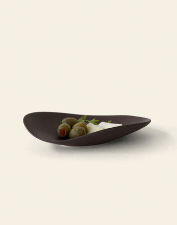 The NR Ceramics HIN Pebble Plate S, a dark, modern piece from NR Ceramics, elegantly holds three green olives and a wedge of white cheese with herbs, set against a light background—perfect tableware for any setting.