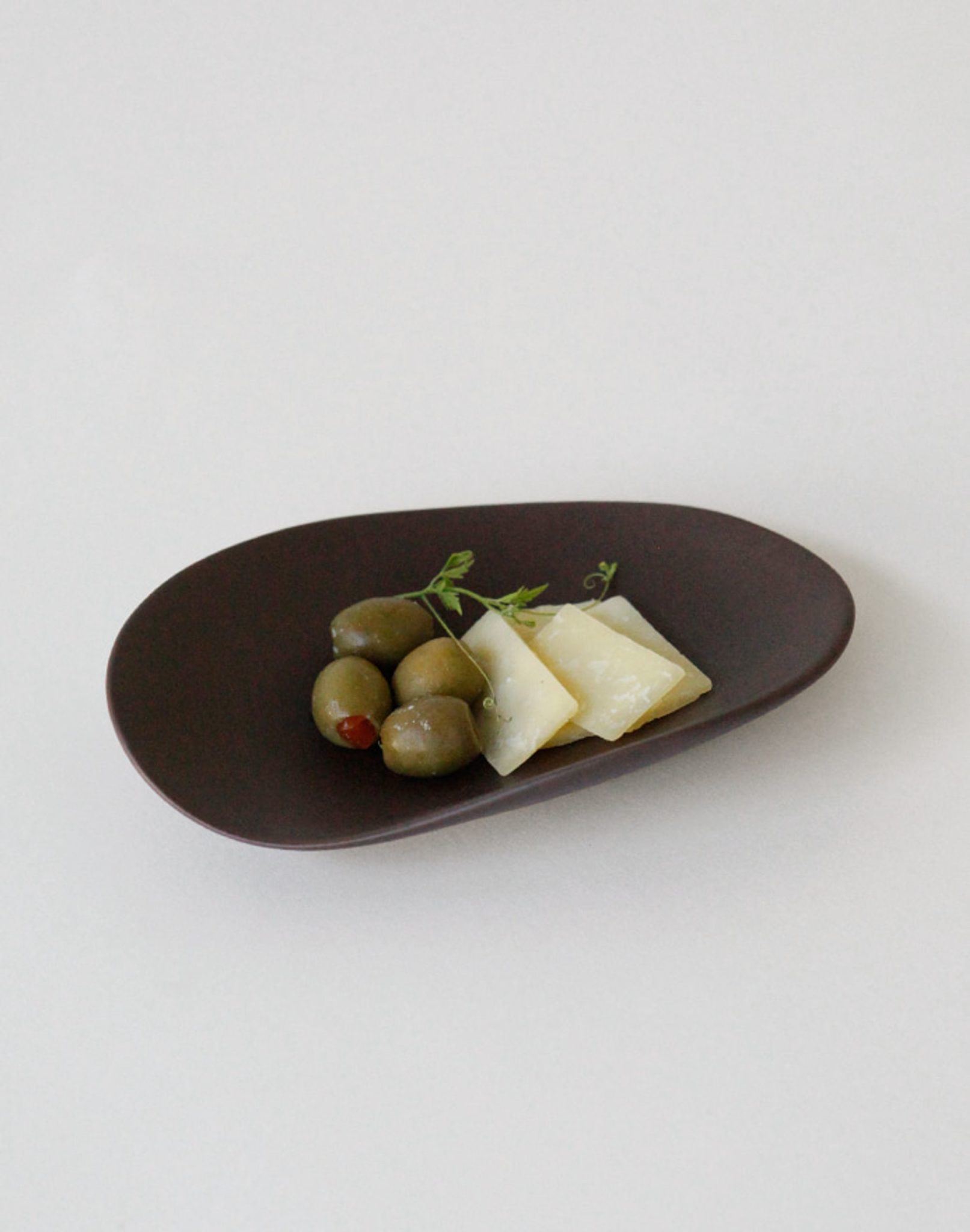 Green olives and two slices of pale yellow cheese, garnished with herbs, are served on the NR Ceramics HIN Pebble Plate S by NR Ceramics—a stylish piece of modern tableware set against a plain white background.