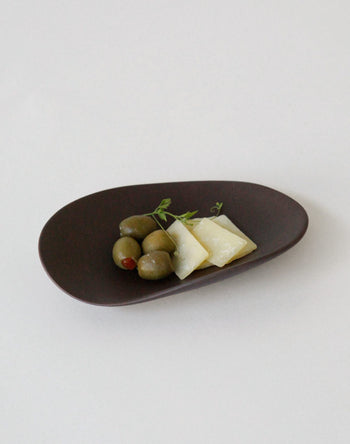 Green olives and two slices of pale yellow cheese, garnished with herbs, are served on the NR Ceramics HIN Pebble Plate S by NR Ceramics—a stylish piece of modern tableware set against a plain white background.