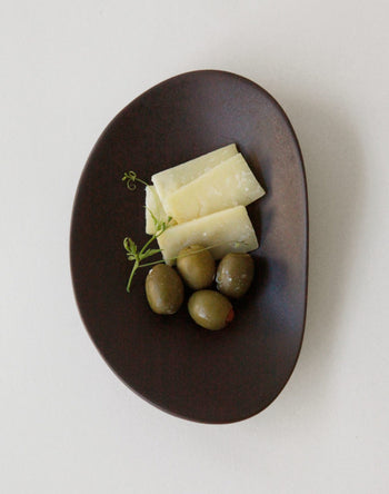 A dark oval NR Ceramics HIN Pebble Plate S by NR Ceramics holds three green olives, three rectangular slices of pale yellow cheese, and a small garnish of fresh herbs on a plain white background.