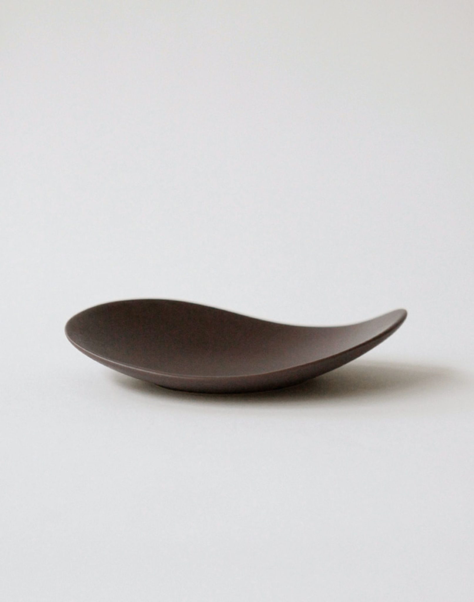 The NR Ceramics HIN Pebble Plate S, a minimalist dark brown ceramic dish from NR Ceramics, features a smooth, wave-like form and sits on a simple light background—an elegant addition to any modern tableware setting.