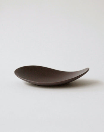 The NR Ceramics HIN Pebble Plate S, a minimalist dark brown ceramic dish from NR Ceramics, features a smooth, wave-like form and sits on a simple light background—an elegant addition to any modern tableware setting.