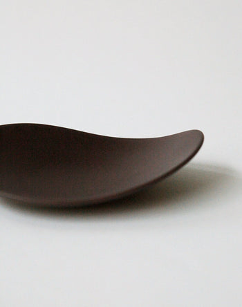 The NR Ceramics HIN Pebble Plate S, a minimalist dark brown piece with a smooth, wave-like shape, is partially visible against a plain white background.