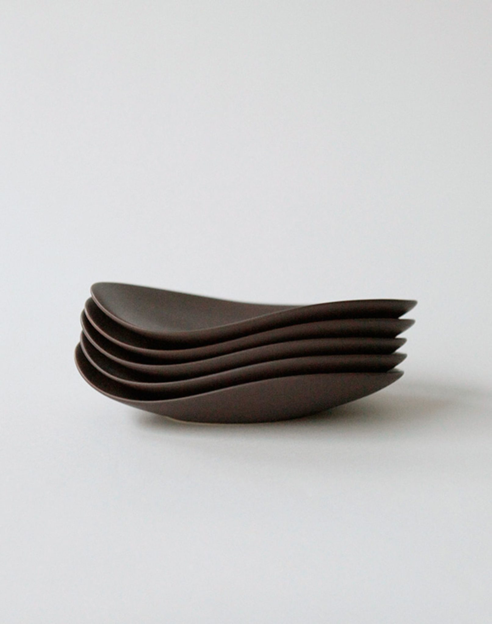 Five NR Ceramics HIN Pebble Plate S in dark brown are stacked neatly on a white surface, their smooth, curved edges showcasing the brand’s modern tableware aesthetic.