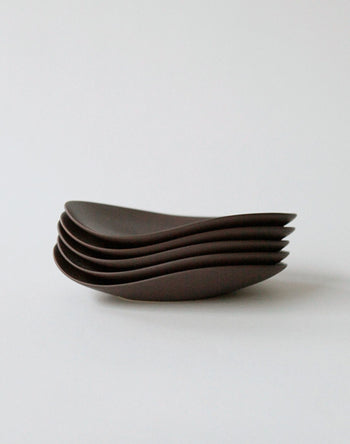 Five NR Ceramics HIN Pebble Plate S in dark brown are stacked neatly on a white surface, their smooth, curved edges showcasing the brand’s modern tableware aesthetic.