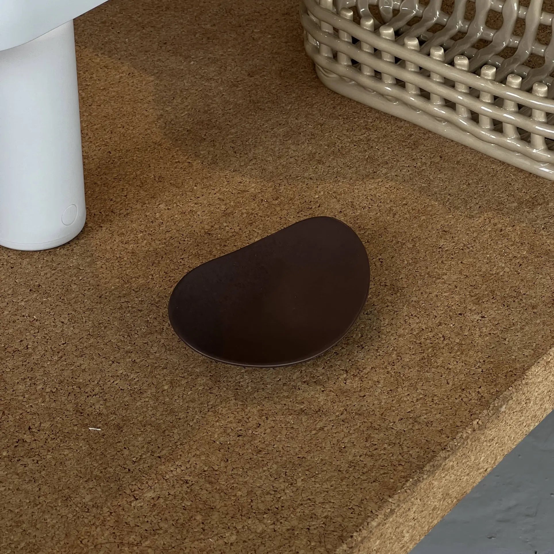 A small, dark brown NR Ceramics HIN Pebble Plate S rests on a cork surface beside a white cylindrical object and a beige woven basket.