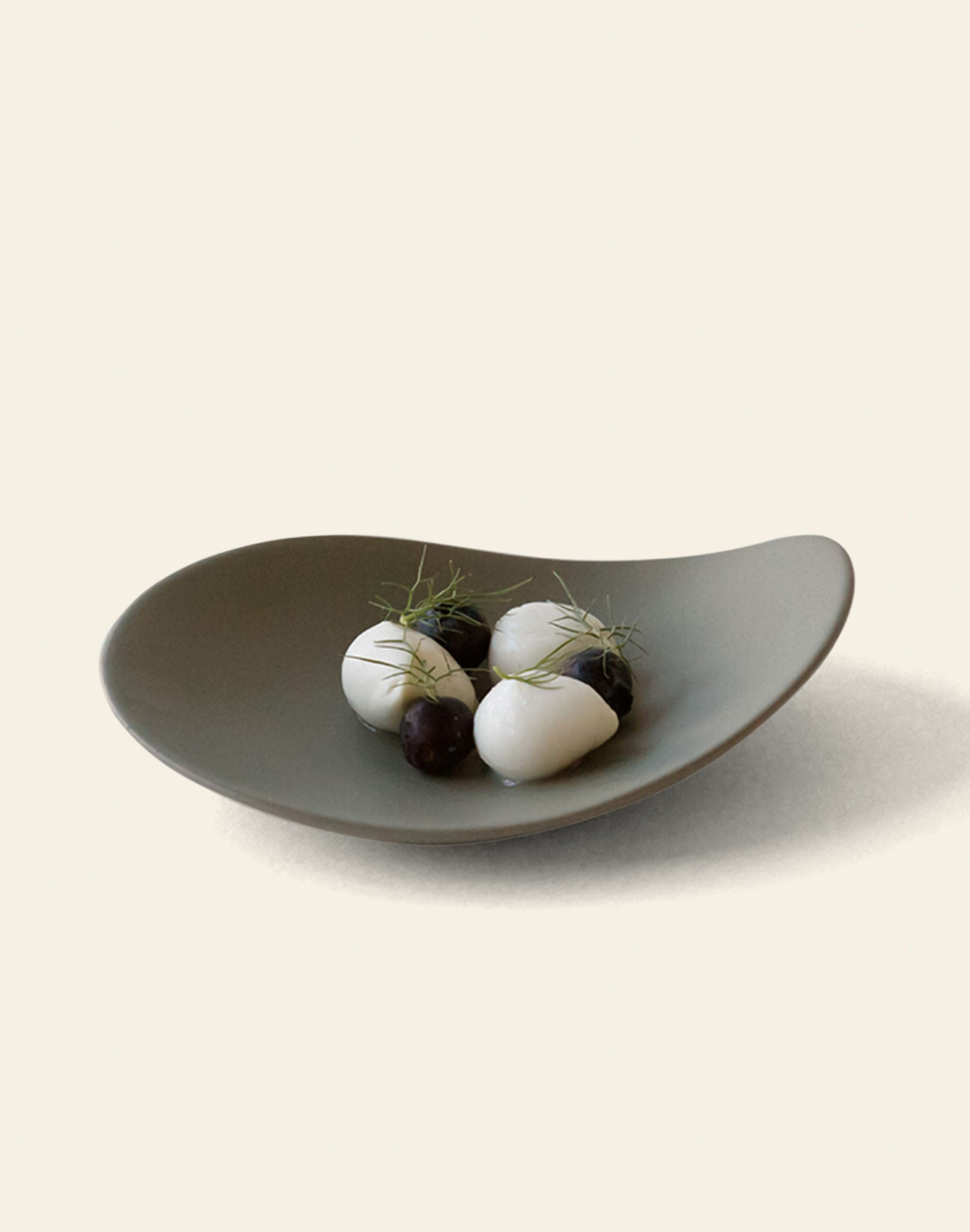 The NR Ceramics HIN Pebble Plate S stylishly holds three white ovals, small dark spheres, and delicate green herbs, displayed on a plain light background. This elegant NR Ceramics piece elevates any dining experience.