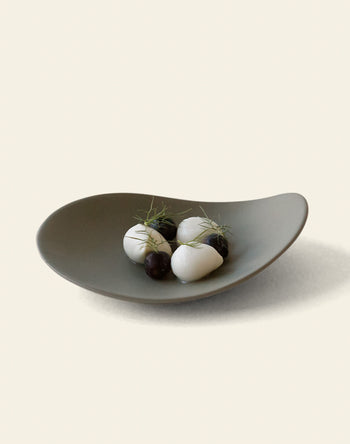 The NR Ceramics HIN Pebble Plate S stylishly holds three white ovals, small dark spheres, and delicate green herbs, displayed on a plain light background. This elegant NR Ceramics piece elevates any dining experience.