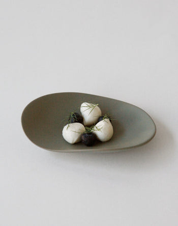 The NR Ceramics HIN Pebble Plate S displays four small white balls with green herbs and two dark balls, arranged on matte, asymmetrical tableware from NR Ceramics, set against a plain white background.