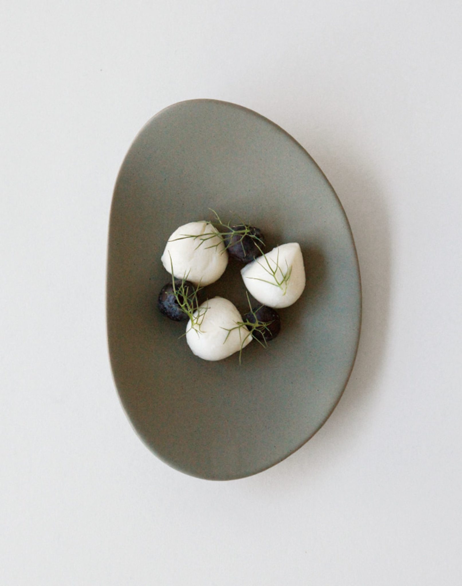 The NR Ceramics HIN Pebble Plate S, in minimalist grey, elegantly displays three mozzarella balls, three blueberries, and green herb sprigs on a plain white background—a perfect example of modern NR Ceramics tableware.