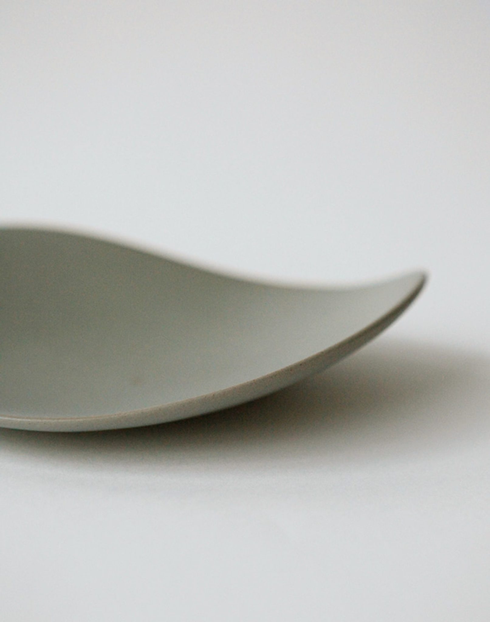 A close-up of the NR Ceramics HIN Pebble Plate S by NR Ceramics is shown on a white background. Only part of this modern plate is visible, highlighting its smooth gray surface and minimalist, curved, wave-like design.