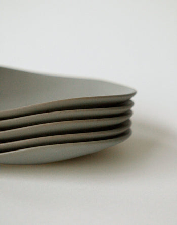 A close-up shows six neatly stacked NR Ceramics HIN Pebble Plate S tableware pieces with subtly wavy edges, displayed on a white surface against a plain background.