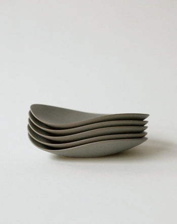 The NR Ceramics HIN Pebble Plate S by NR Ceramics rests on a plain white surface, its minimalist gray ceramic form creating a smooth, curved silhouette that elevates modern tableware.
