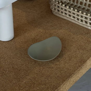 The NR Ceramics HIN Pebble Plate S—a small, matte gray plate with an irregular shape—rests on a cork tabletop near a white cylinder and beige woven basket, bringing charm to modern tableware arrangements.