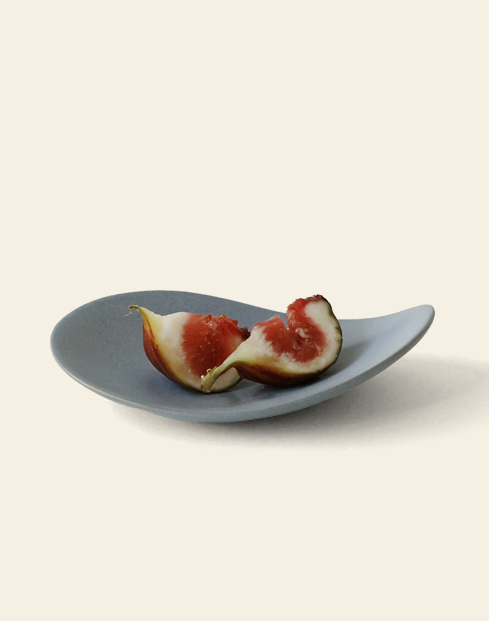A halved ripe fig with red flesh rests on the NR Ceramics HIN Pebble Plate S, an asymmetrical gray plate by NR Ceramics, displayed against a plain light background.