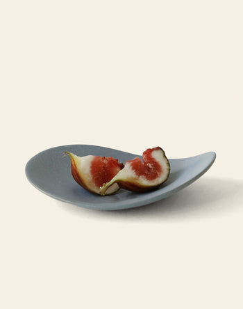 A halved ripe fig with red flesh rests on the NR Ceramics HIN Pebble Plate S, an asymmetrical gray plate by NR Ceramics, displayed against a plain light background.