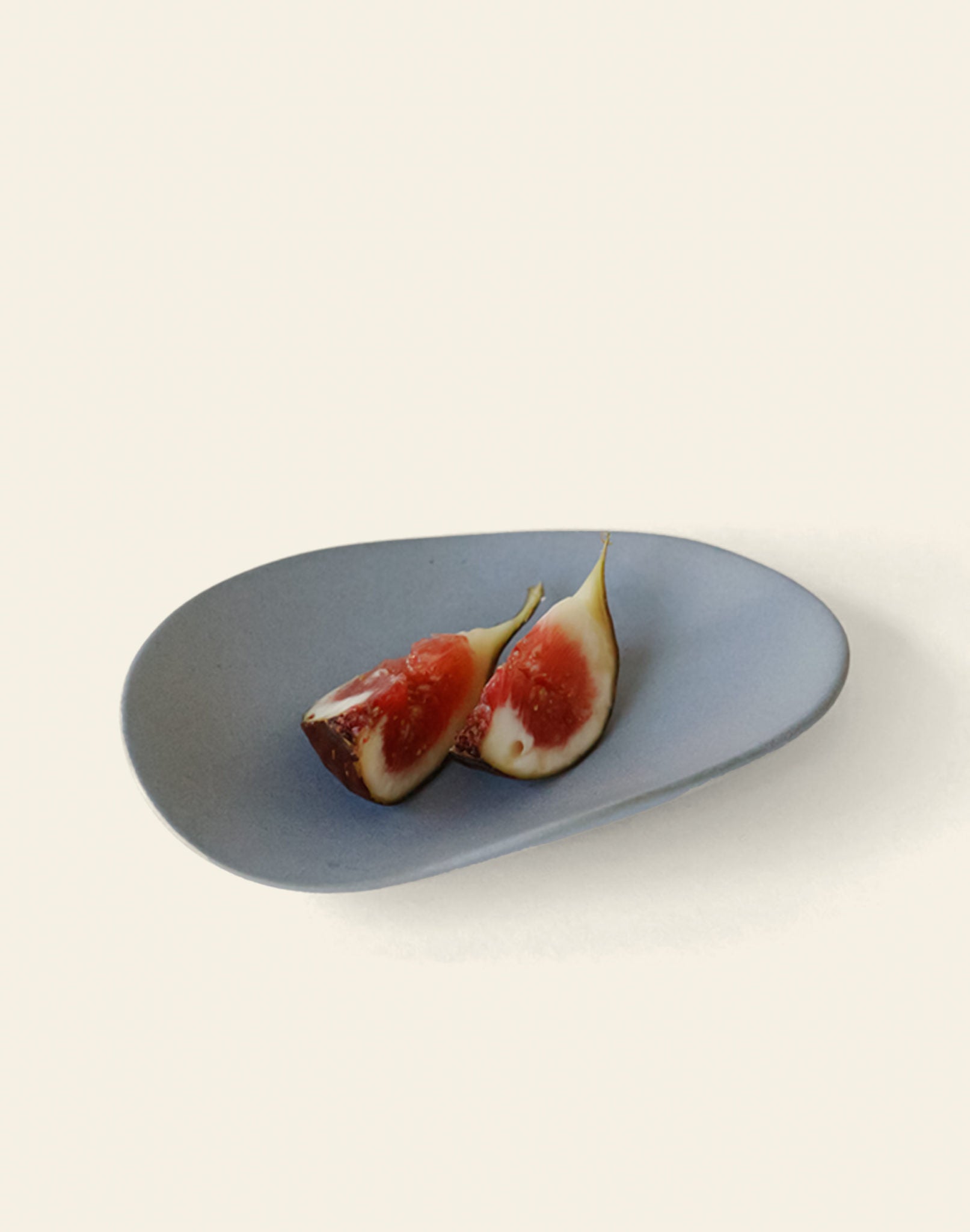 A minimalist photo shows two fig halves with pink flesh on the NR Ceramics HIN Pebble Plate S by NR Ceramics, set against a simple, light background.