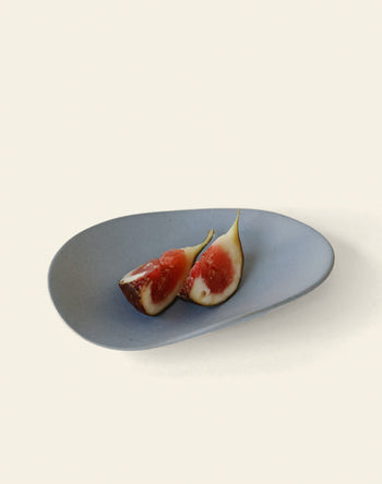 A minimalist photo shows two fig halves with pink flesh on the NR Ceramics HIN Pebble Plate S by NR Ceramics, set against a simple, light background.