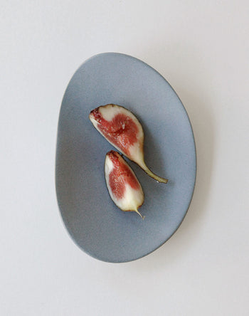 Two halves of a fresh fig with a reddish interior rest on the NR Ceramics HIN Pebble Plate S from NR Ceramics, displayed on a smooth, oval surface against a white background.