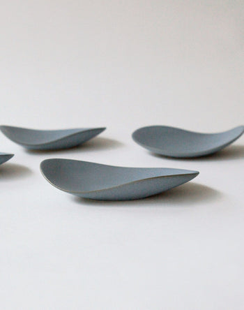 Four minimalist NR Ceramics HIN Pebble Plate S dishes are arranged on a plain white surface, each with a smooth, modern design resembling gently upturned leaves or pebbles.