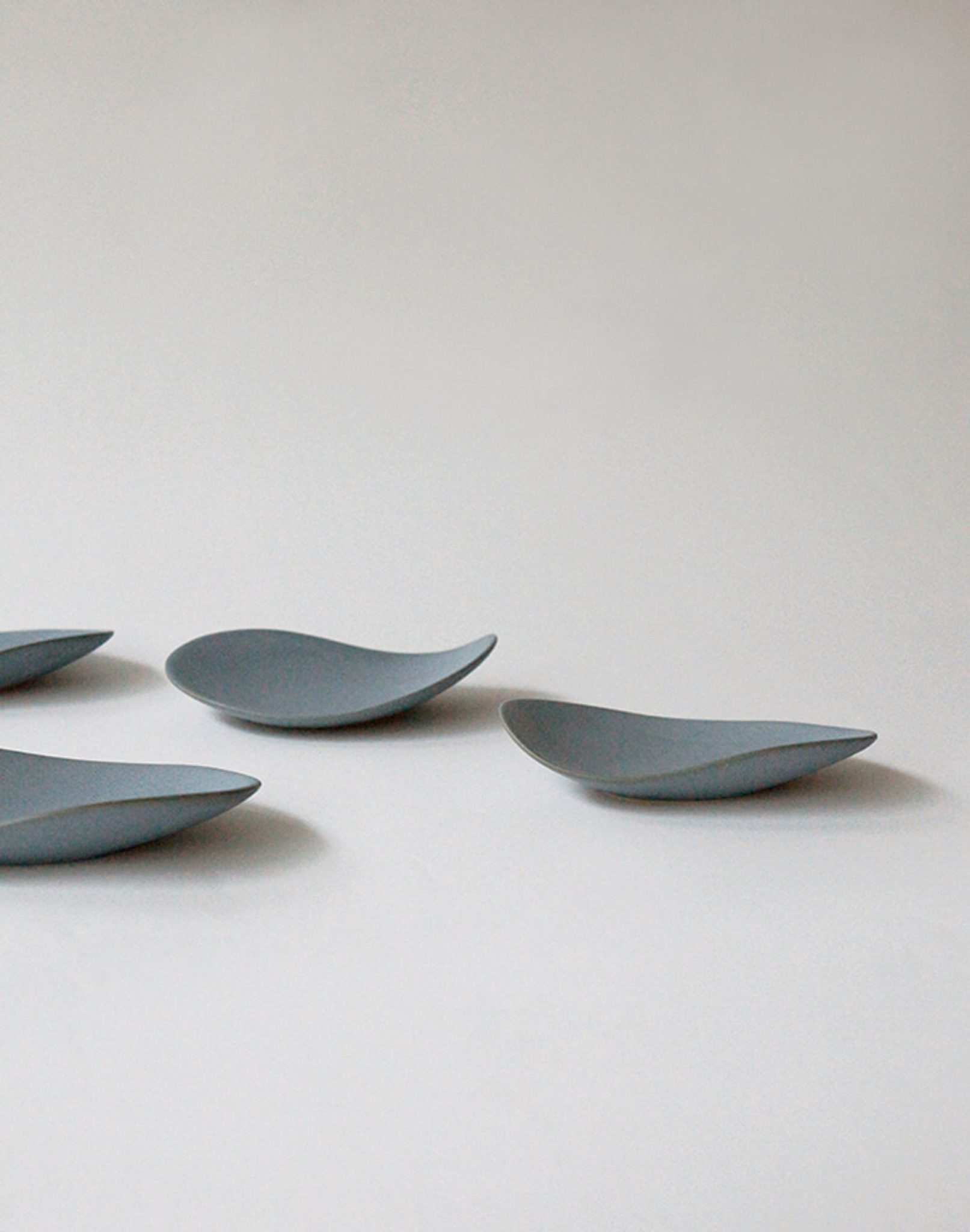 Four NR Ceramics HIN Pebble Plate S dishes, in gray with smooth, organic curves, are arranged on a light surface with ample negative space, showcasing the minimalist elegance of NR Ceramics modern tableware.
