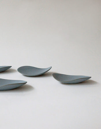 Four NR Ceramics HIN Pebble Plate S dishes, in gray with smooth, organic curves, are arranged on a light surface with ample negative space, showcasing the minimalist elegance of NR Ceramics modern tableware.