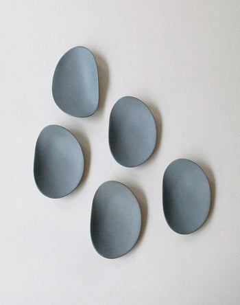 Five smooth, gray NR Ceramics HIN Pebble Plate S are arranged on a light surface in a loose circle, evoking flat stones—an elegant, contemporary tableware accent from NR Ceramics.