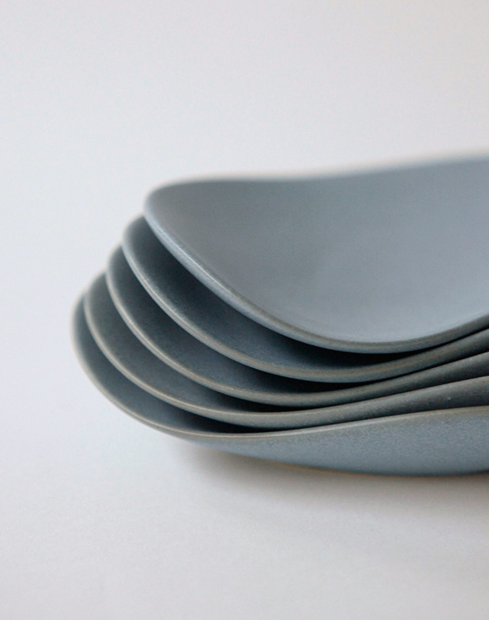 A close-up of five NR Ceramics HIN Pebble Plate S dishes, neatly stacked in a fanned-out formation on a light surface—ideal for adding minimalist elegance to modern table settings.