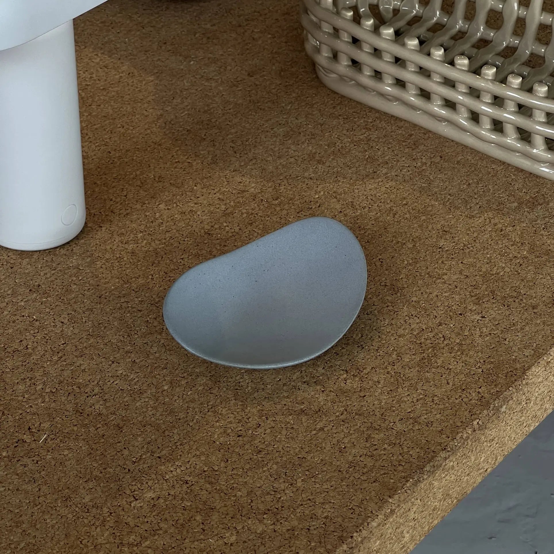 The NR Ceramics HIN Pebble Plate S, a small asymmetrical gray plate, rests on a cork surface beside a white cylindrical item and a beige woven basket.