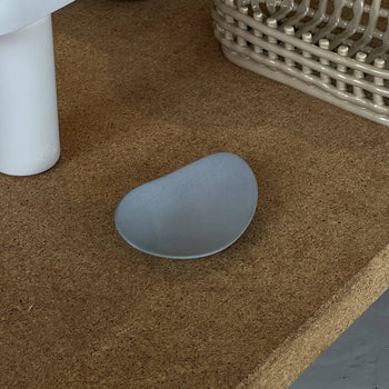 The NR Ceramics HIN Pebble Plate S, a small asymmetrical gray plate, rests on a cork surface beside a white cylindrical item and a beige woven basket.