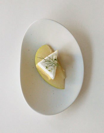 A small wedge of cheese with a white rind rests on green apple, garnished with herbs, and is elegantly served on the NR Ceramics HIN Pebble Plate S by NR Ceramics, set against a plain white background.