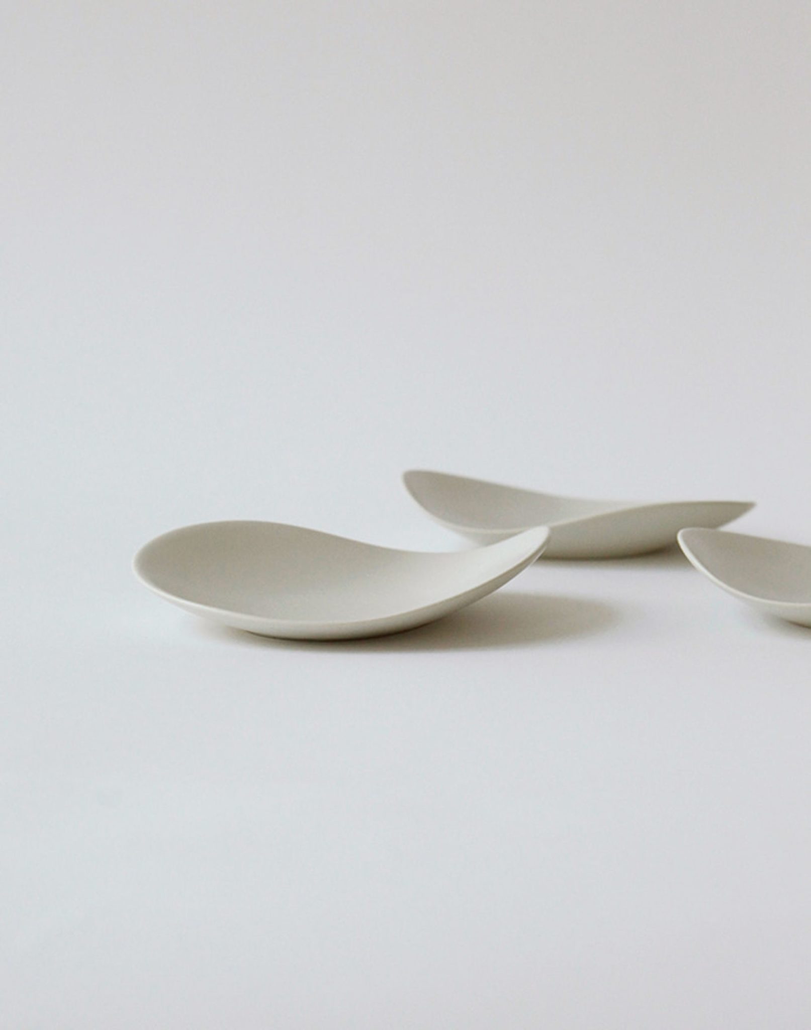 Three minimalist, curved white NR Ceramics HIN Pebble Plate S dishes are arranged on a plain white surface, forming a clean and modern tableware display with soft shadows.
