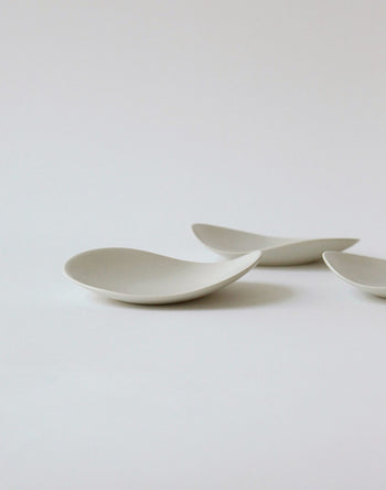 Three minimalist, curved white NR Ceramics HIN Pebble Plate S dishes are arranged on a plain white surface, forming a clean and modern tableware display with soft shadows.