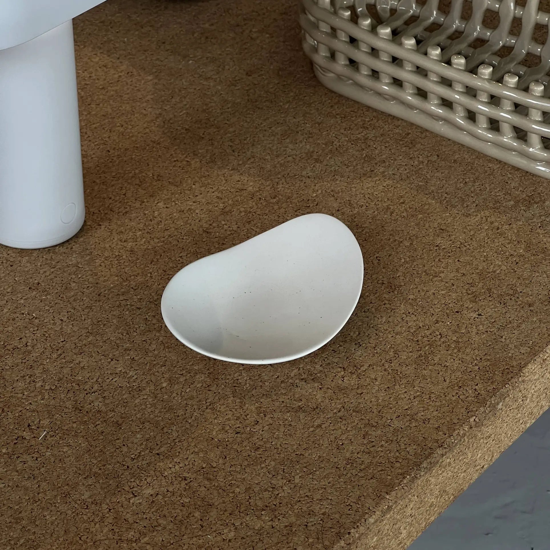 The NR Ceramics HIN Pebble Plate S, a small irregular white plate, rests on cork near a white cylinder and beige basket, bringing charm to modern table settings.