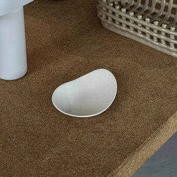 The NR Ceramics HIN Pebble Plate S, a small irregular white plate, rests on cork near a white cylinder and beige basket, bringing charm to modern table settings.