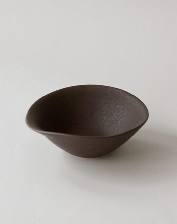 The NR Ceramics HIN Soup Bowl features a dark brown, asymmetrical ceramic design. Displayed on a plain, light surface with soft lighting, this versatile tableware by NR Ceramics suits any setting.
