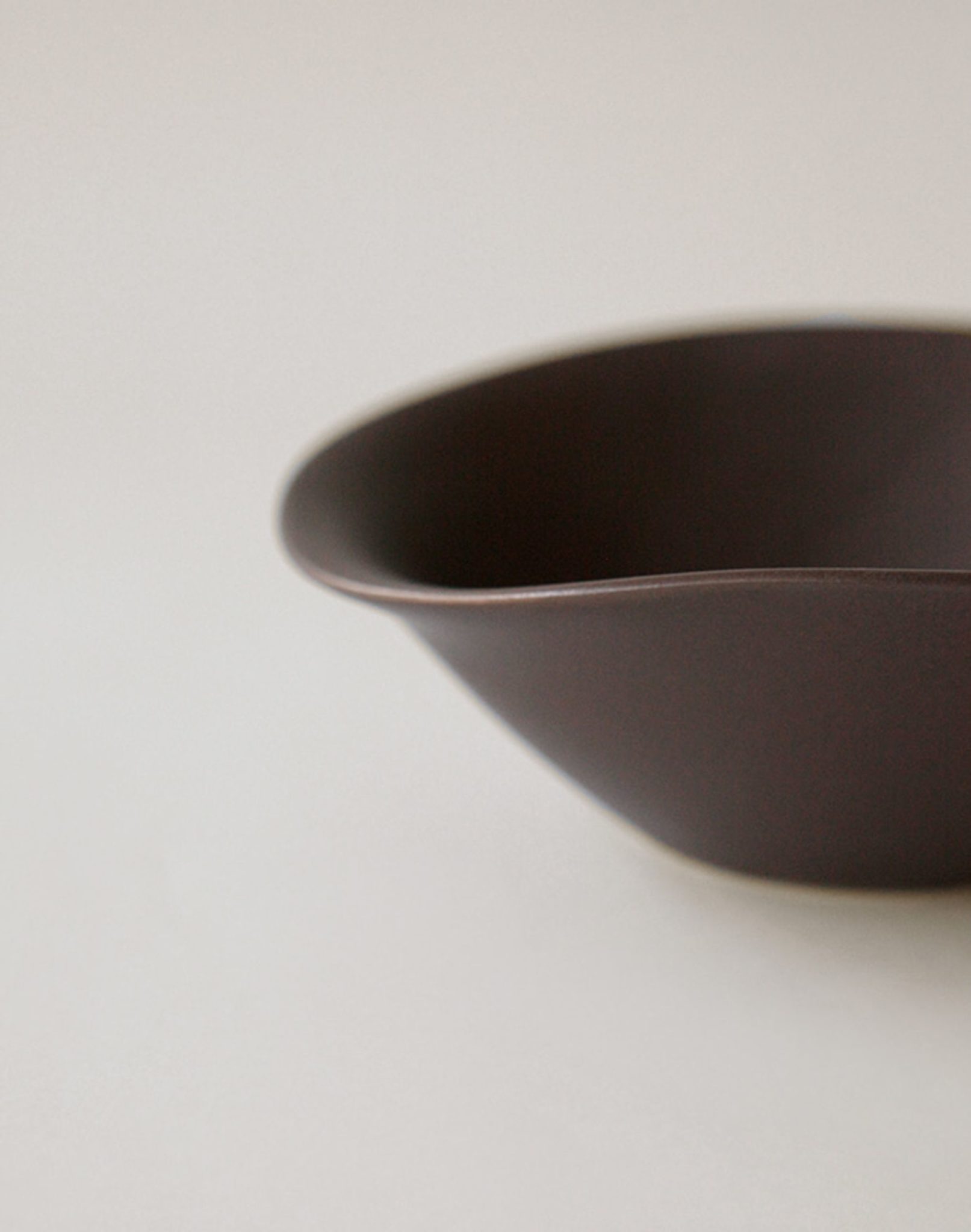 A close-up of the NR Ceramics HIN Soup Bowl, a smooth, dark brown ceramic piece with soft curves from NR Ceramics, set on a light surface against a plain neutral background.