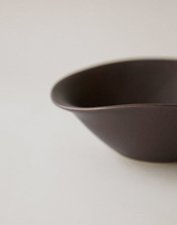 A close-up of the NR Ceramics HIN Soup Bowl, a smooth, dark brown ceramic piece with soft curves from NR Ceramics, set on a light surface against a plain neutral background.